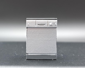 Könnte beinhalten: Ein kleiner, grauer Miniatur-Geschirrspüler mit strukturierter Oberfläche. Das Gerät hat einen Griff und Details des Bedienfelds. Die Miniatur befindet sich vor einem dunklen Hintergrund.