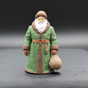 Può includere: Una statuetta di Babbo Natale con un cappotto verde con finiture marroni e un cappello abbinato. La figura ha una barba bianca e tiene in mano un sacco beige. Il cappotto ha un motivo decorativo e una cintura marrone.
