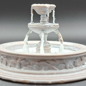 May include: A miniature fountain with a tiered design, featuring a central spout and water cascading into a round basin. The fountain has a weathered, antique appearance with a light brown and white colour scheme. The basin is decorated with relief carvings.