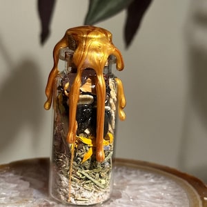 May include: A small, clear glass bottle filled with dried herbs, flowers, and crystals. The bottle is sealed with a golden, dripped wax. The bottle is sitting on a light brown and white stone.