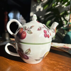 May include: A white porcelain teacup and teapot set with a fruit design. The teapot sits atop the teacup, featuring illustrations of red apples, plums, and cherries with green leaves. A green band encircles the teacup and the lid of the teapot.