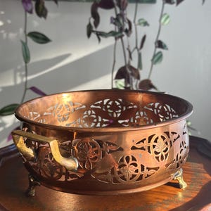 May include: A copper-colored bowl with intricate cut-out floral designs. The bowl has two brass handles and rests on small brass feet. The bowl is sitting on a wooden table, with a plant in the background.
