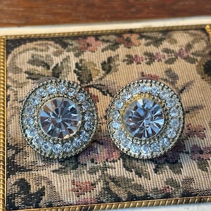 May include: A pair of vintage-style stud earrings. Each earring features a large, clear, faceted crystal center stone surrounded by a halo of smaller, clear crystals. The earrings have a gold-tone metal setting.