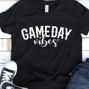 K&ouml;nnte beinhalten: Schwarzes T-Shirt mit wei&szlig;em Text, der "GAMEDAY vibes" mit einem kleinen Herzsymbol lautet. Das Shirt liegt flach auf einer wei&szlig;en Holzoberfl&auml;che mit einem Paar schwarzer Sneakers und einer blauen Jeans.