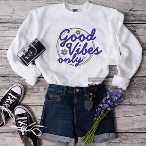 Pode incluir: Camisola branca com um gr&aacute;fico circular castanho que diz "Good Vibes Only" em texto azul com um cora&ccedil;&atilde;o rosa. A camisola est&aacute; colocada sobre um fundo de madeira com um par de cal&ccedil;&otilde;es de ganga azul e um par de sapatos de lona pretos.