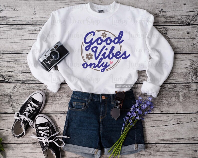 Pode incluir: Camisola branca com um gr&aacute;fico circular castanho que diz "Good Vibes Only" em texto azul com um cora&ccedil;&atilde;o rosa. A camisola est&aacute; colocada sobre um fundo de madeira com um par de cal&ccedil;&otilde;es de ganga azul e um par de sapatos de lona pretos.