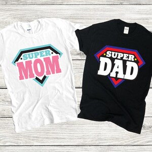 May include: Two t-shirts with a superhero theme. One white t-shirt says "SUPER MOM" in a pink, black, and blue diamond shape. The other black t-shirt says "SUPER DAD" in a red, blue, and white diamond shape.