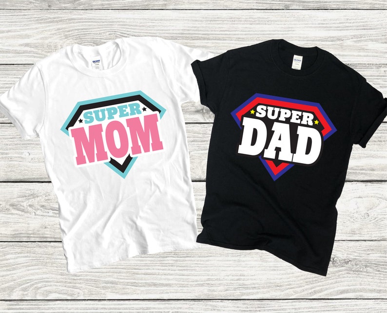 May include: Two t-shirts with a superhero theme. One white t-shirt says "SUPER MOM" in a pink, black, and blue diamond shape. The other black t-shirt says "SUPER DAD" in a red, blue, and white diamond shape.