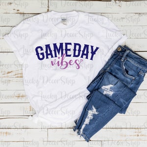 K&ouml;nnte beinhalten: Wei&szlig;es T-Shirt mit dem Text "GAMEDAY vibes" in blauer und rosa Schrift auf wei&szlig;em Holzgrund. Eine blaue Jeans ist auf dem T-Shirt gefaltet.