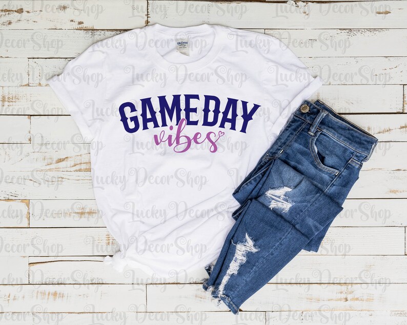 K&ouml;nnte beinhalten: Wei&szlig;es T-Shirt mit dem Text "GAMEDAY vibes" in blauer und rosa Schrift auf wei&szlig;em Holzgrund. Eine blaue Jeans ist auf dem T-Shirt gefaltet.