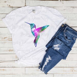 Puede incluir: Una camiseta blanca con un gr&aacute;fico de colibr&iacute; colorido. El colibr&iacute; es de estilo acuarela con colores rosa, azul, verde y morado. La camiseta est&aacute; sobre un fondo de madera blanca con un par de pantalones vaqueros azules.