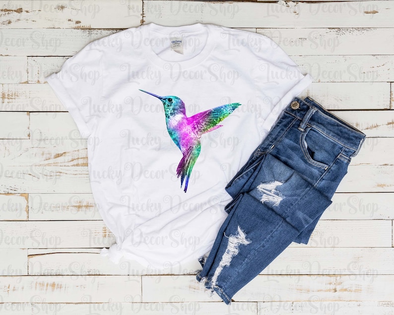 Puede incluir: Una camiseta blanca con un gr&aacute;fico de colibr&iacute; colorido. El colibr&iacute; es de estilo acuarela con colores rosa, azul, verde y morado. La camiseta est&aacute; sobre un fondo de madera blanca con un par de pantalones vaqueros azules.