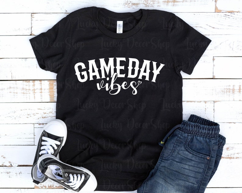 K&ouml;nnte beinhalten: Schwarzes T-Shirt mit wei&szlig;em Text, der "GAMEDAY vibes" mit einem kleinen Herzsymbol lautet. Das Shirt liegt flach auf einer wei&szlig;en Holzoberfl&auml;che mit einem Paar schwarzer Sneakers und einer blauen Jeans.