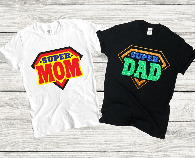 May include: Two t-shirts, one white and one black, with a superhero graphic and the text "Super Mom" and "Super Dad" in red and green glitter.