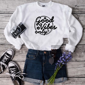 Pode incluir: Camisola branca com texto preto que diz "Good Vibes Only" em um c&iacute;rculo com uma estrela e um cora&ccedil;&atilde;o. A camisola est&aacute; colocada sobre uma superf&iacute;cie de madeira com um par de t&ecirc;nis pretos, shorts jeans azuis e um buqu&ecirc; de flores roxas.