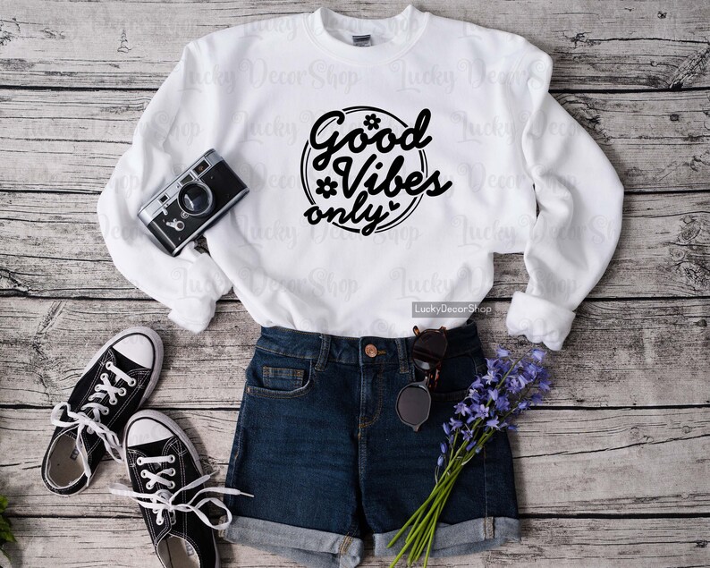 Pode incluir: Camisola branca com texto preto que diz "Good Vibes Only" em um c&iacute;rculo com uma estrela e um cora&ccedil;&atilde;o. A camisola est&aacute; colocada sobre uma superf&iacute;cie de madeira com um par de t&ecirc;nis pretos, shorts jeans azuis e um buqu&ecirc; de flores roxas.
