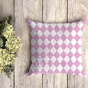 May include: A pink and white diamond patterned cushion. The cushion is on a light wood background.