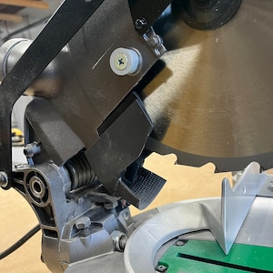 Könnte beinhalten: Nahaufnahme einer schwarzen und silbernen Gehrungssäge mit einem Metallblatt. Die Säge ist positioniert, um ein Stück Holz zu schneiden.