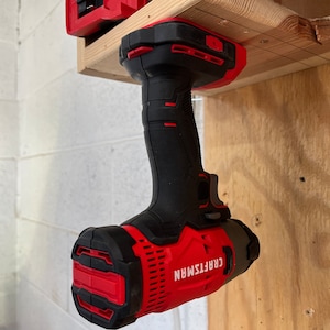 May include: A red and black cordless drill with the brand name "CRAFTSMAN" printed on the side. The drill is hanging upside down from a wooden shelf.