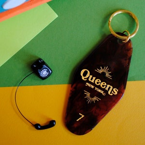 May include: A brown motel key chain with gold lettering that reads "Queens New York" and the number "7". The key chain is on a green and yellow background with a small black rotary phone.