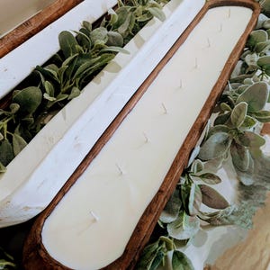 Pu&ograve; includere: Una lunga candela rettangolare in una vasca di legno con pi&ugrave; stoppini. La candela &egrave; bianca e la vasca &egrave; di colore marrone scuro. La candela &egrave; circondata da vegetazione, creando un'estetica rustica e naturale.