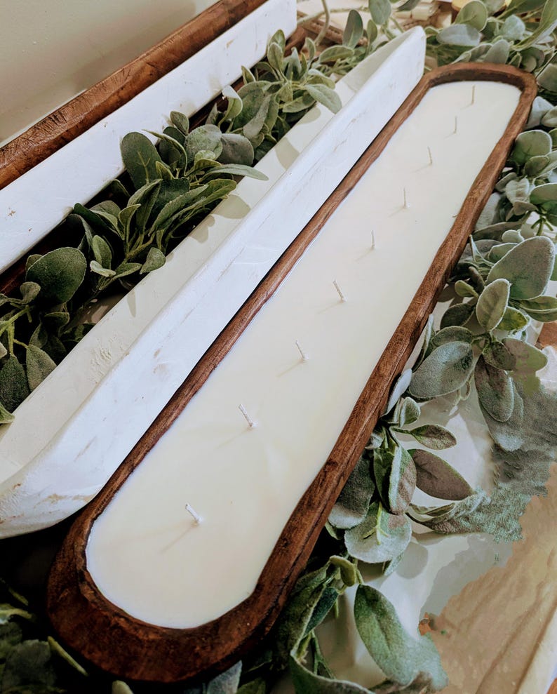 Pu&ograve; includere: Una lunga candela rettangolare in una vasca di legno con pi&ugrave; stoppini. La candela &egrave; bianca e la vasca &egrave; di colore marrone scuro. La candela &egrave; circondata da vegetazione, creando un'estetica rustica e naturale.