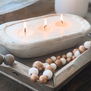 May include: A white, oval-shaped dough bowl candle featuring three lit wicks and smooth white wax. The candle rests on a rectangular wooden tray decorated with a string of alternating metallic gold and matte white wooden beads.