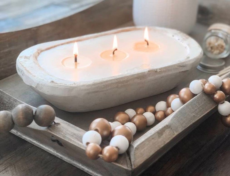 May include: A white, oval-shaped dough bowl candle featuring three lit wicks and smooth white wax. The candle rests on a rectangular wooden tray decorated with a string of alternating metallic gold and matte white wooden beads.