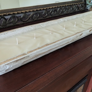 Pu&ograve; includere: Una lunga candela rettangolare in una vasca di legno bianco anticato. La candela ha cera bianco crema e stoppini multipli. La vasca &egrave; posizionata su una superficie marrone scuro, con uno specchio decorativo sullo sfondo. La candela &egrave; un oggetto di arredamento.