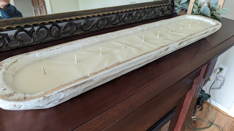 Pu&ograve; includere: Una lunga candela rettangolare in una vasca di legno bianco anticato. La candela ha cera bianco crema e stoppini multipli. La vasca &egrave; posizionata su una superficie marrone scuro, con uno specchio decorativo sullo sfondo. La candela &egrave; un oggetto di arredamento.