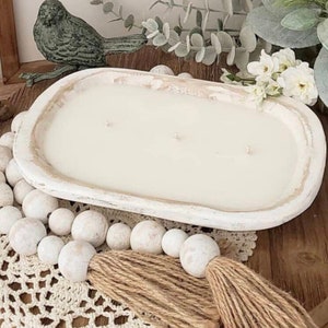 May include: An oval-shaped, distressed white wooden dough bowl candle featuring three wicks and smooth white wax. The bowl is accented by a strand of white wooden beads with a jute tassel, positioned on a white crocheted lace mat.