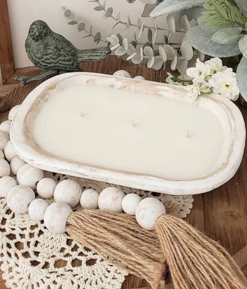 May include: An oval-shaped, distressed white wooden dough bowl candle featuring three wicks and smooth white wax. The bowl is accented by a strand of white wooden beads with a jute tassel, positioned on a white crocheted lace mat.