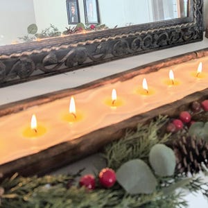 Pu&ograve; includere: Un lungo portacandele in legno con pi&ugrave; candele accese. Le candele sono poste in una rustica vasca di legno intagliato, circondata da verde, pigne e bacche rosse. Le candele sono illuminate da un bagliore caldo.