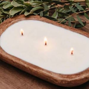 May include: A three-wick candle set in an oval-shaped, rustic brown wooden dough bowl. The candle features smooth, white wax with three small, lit flames positioned along the centre. The bowl has a textured, natural wood grain finish.