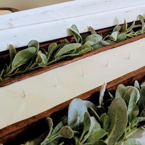 Pu&ograve; includere: Un grande portacandele rettangolare in legno riempito di cera color crema e stoppini multipli. Il portacandele &egrave; circondato da fogliame verde e ci sono vassoi di legno bianchi aggiuntivi sullo sfondo. Un oggetto di arredamento rustico.