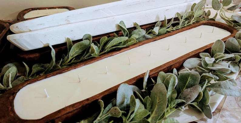 Pu&ograve; includere: Un grande portacandele rettangolare in legno riempito di cera color crema e stoppini multipli. Il portacandele &egrave; circondato da fogliame verde e ci sono vassoi di legno bianchi aggiuntivi sullo sfondo. Un oggetto di arredamento rustico.