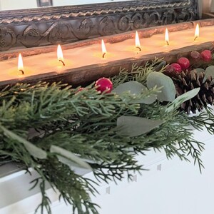 Pu&ograve; includere: Un lungo portacandele rettangolare in legno con pi&ugrave; stoppini accesi. Il portacandele &egrave; decorato con pigne, bacche rosse e rami sempreverdi. Le candele bruciano con un bagliore caldo e invitante, creando un'atmosfera festosa.