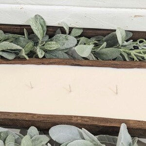 Pu&ograve; includere: Una candela lunga e rettangolare con pi&ugrave; stoppini, posta in una rustica vasca di legno marrone scuro. La candela ha una cera bianco crema ed &egrave; circondata da fogliame verde e grigio, creando un'estetica naturale da fattoria.