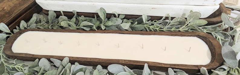 Pu&ograve; includere: Una candela lunga e rettangolare con pi&ugrave; stoppini, posta in una rustica vasca di legno marrone scuro. La candela ha una cera bianco crema ed &egrave; circondata da fogliame verde e grigio, creando un'estetica naturale da fattoria.