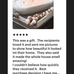 May include: A white, oval-shaped wooden dough bowl candle with three lit wicks, accompanied by a strand of decorative wooden beads in alternating metallic gold and matte white finishes, resting on a wooden tray surface.