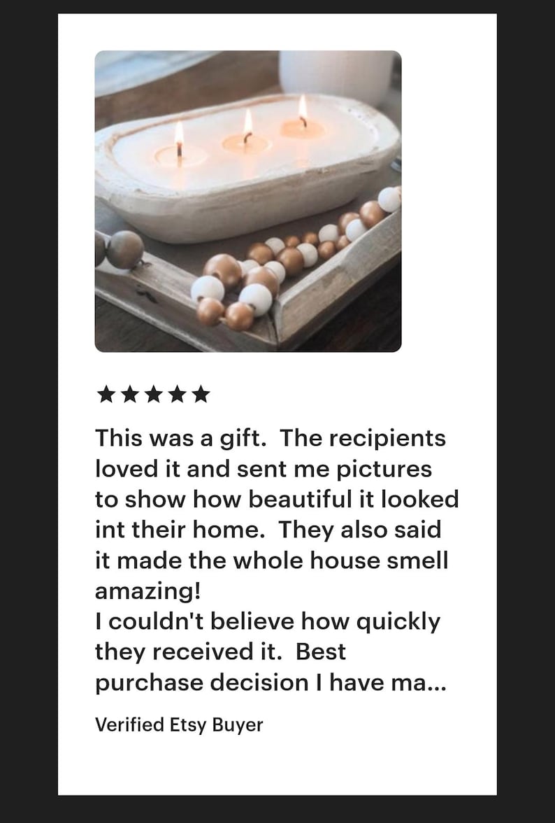 May include: A white, oval-shaped wooden dough bowl candle with three lit wicks, accompanied by a strand of decorative wooden beads in alternating metallic gold and matte white finishes, resting on a wooden tray surface.