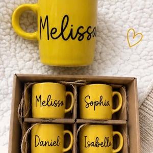 May include: A large yellow ceramic mug with the name Melissa in black script, accompanied by a cardboard gift box containing four smaller yellow ceramic mugs, each personalized with the names Melissa, Sophia, Daniel, and Isabella in black script.