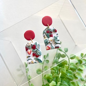 Puede incluir: Un par de pendientes hechos a mano de arcilla polimérica con forma de arco blanco con un patrón de fresas rojas y hojas verdes. Los pendientes tienen tapas circulares rojas.