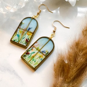 May include: A pair of gold dangle earrings with a window-shaped design featuring a watercolor painting of a field of flowers.