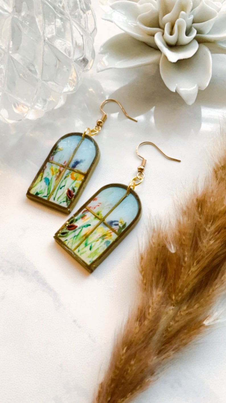 May include: A pair of gold dangle earrings with a window-shaped design featuring a watercolor painting of a field of flowers.