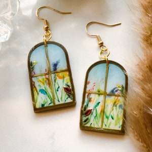 May include: A pair of gold earrings with a window-shaped design featuring a watercolor painting of a garden with colorful flowers.