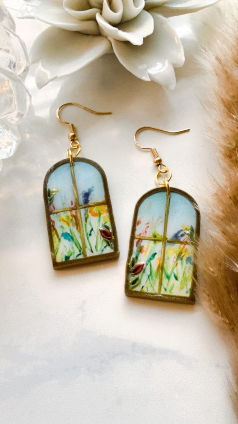 May include: A pair of gold earrings with a window-shaped design featuring a watercolor painting of a garden with colorful flowers.