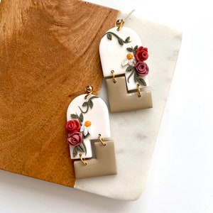 May include: A pair of white and beige polymer clay earrings with a floral design. The earrings feature a white arch with a vine and red and white flowers, and a beige square shape.