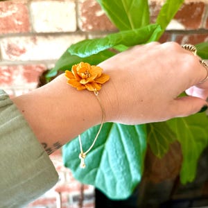Puede incluir: Una pulsera de cadena dorada con un dije de flor naranja detallada. La flor tiene múltiples pétalos y un centro texturizado. La pulsera se lleva en la muñeca, con una manga de camisa verde visible. El fondo incluye una pared de ladrillo y una planta verde.