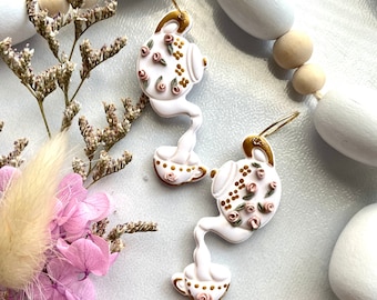 May include: A pair of white teapot earrings with gold accents and pink floral details. The teapots are pouring into a teacup.